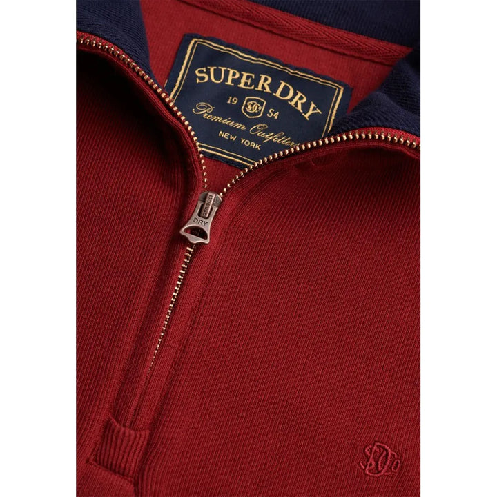 Close-up of burgundy half-zip top: SUPERDRY label, zip with branded pull tab