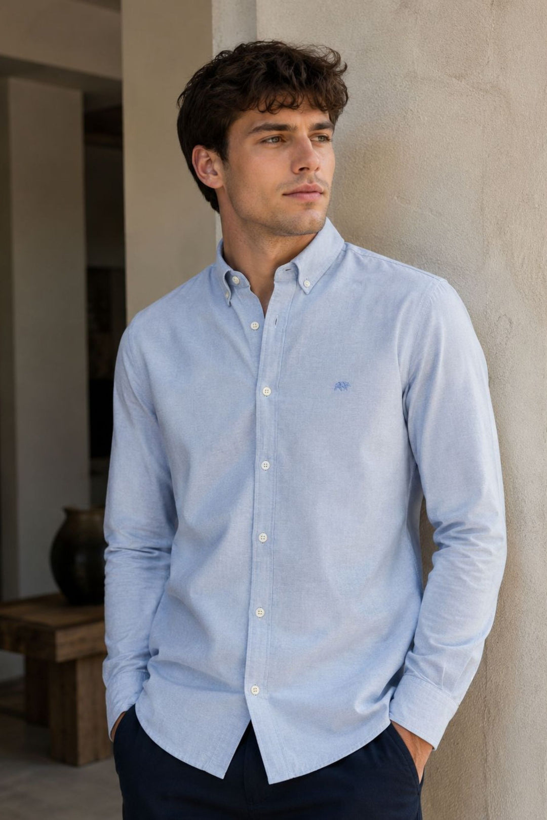 Man wearing a light blue Rivet & Denim Oxford shirt standing against a neutral wall.